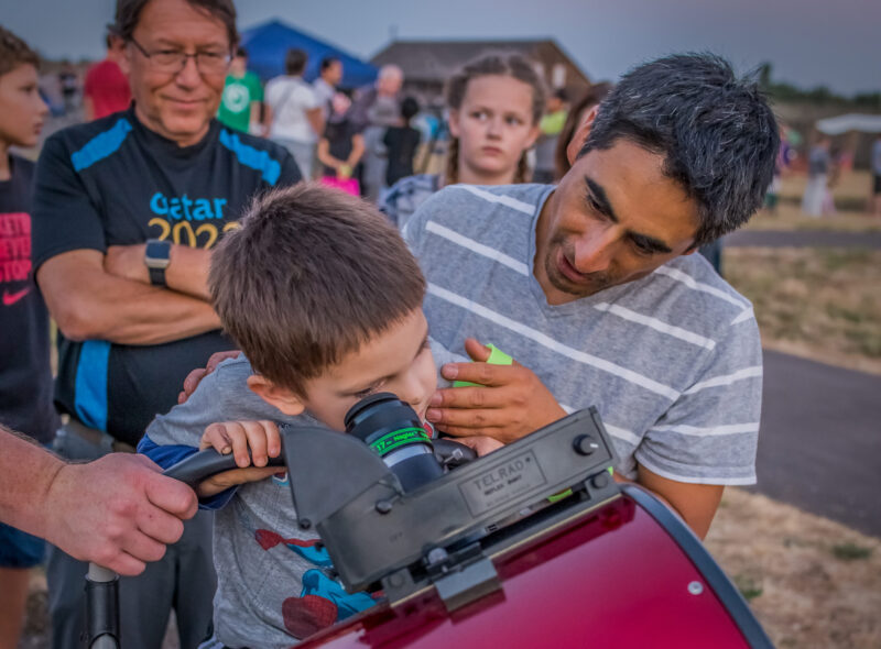 Child looks at the sky through a large telescope with the help of an adult.