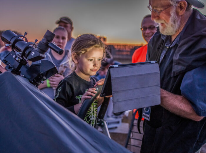 Child looks at a star map on an iPad next to a large telescope