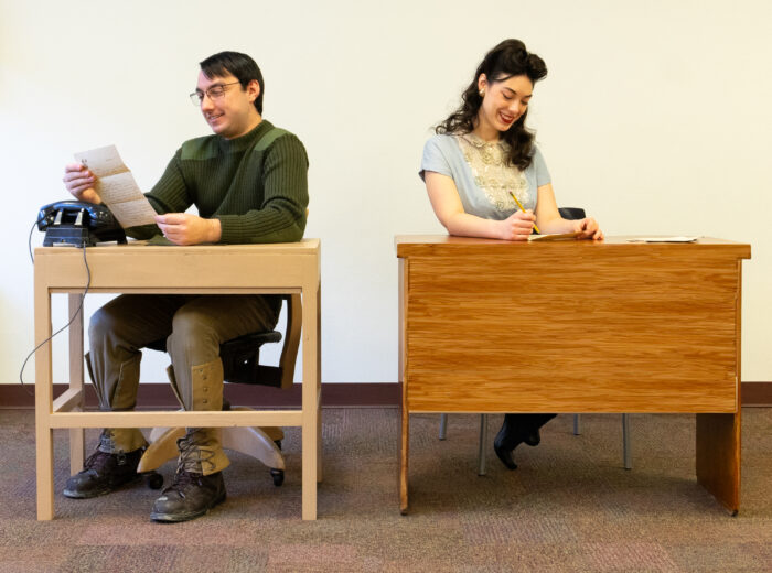 Two actors sit at different desks, writing letters.